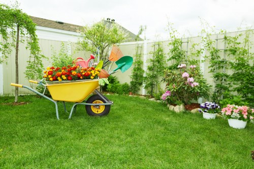 A well-maintained backyard garden with a lush, green lawn occupying most of the space, bordered by a white wooden fence with vertical panels and supporting climbing plants and greenery. In the foreground, a yellow wheelbarrow filled with colorful flowers and garden tools is positioned on the grass, indicating recent gardening activity. Along the right side, there are flower beds containing blossoming pink and purple flowers, arranged with a mix of soil and mulch surrounding the plants. The background features small trees and shrubs, creating a natural border and adding layer to the garden environment. The overall scene is outdoors with natural daylight, suggesting a bright, partly cloudy day typical of spring or summer, and it reflects a tidy, inviting space suitable for garden maintenance and landscaping services provided by Gardener Bounds Green.